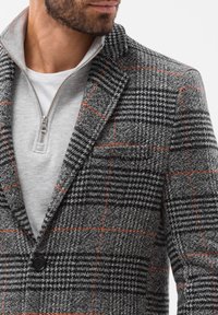 Gråternet blazer med orange detaljer; har en klapkrave og en enkelt knap, lagdelt over en grå lynlåstrøje.