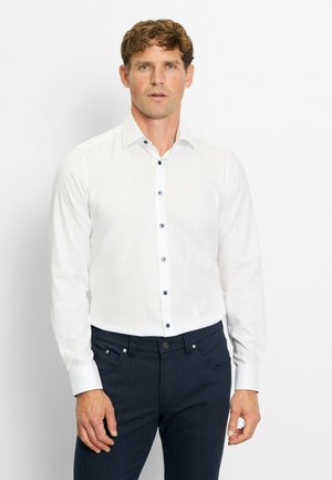 White long-sleeve shirt with a button-down collar and navy buttons, paired with dark blue trousers. Smooth fabric, tailored fit.