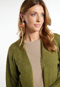 Cardigan verde oliva con apertura frontale, texture morbida; abbinato a una maglietta beige. Presenta grandi orecchini rotondi colorati con accenti metallici.