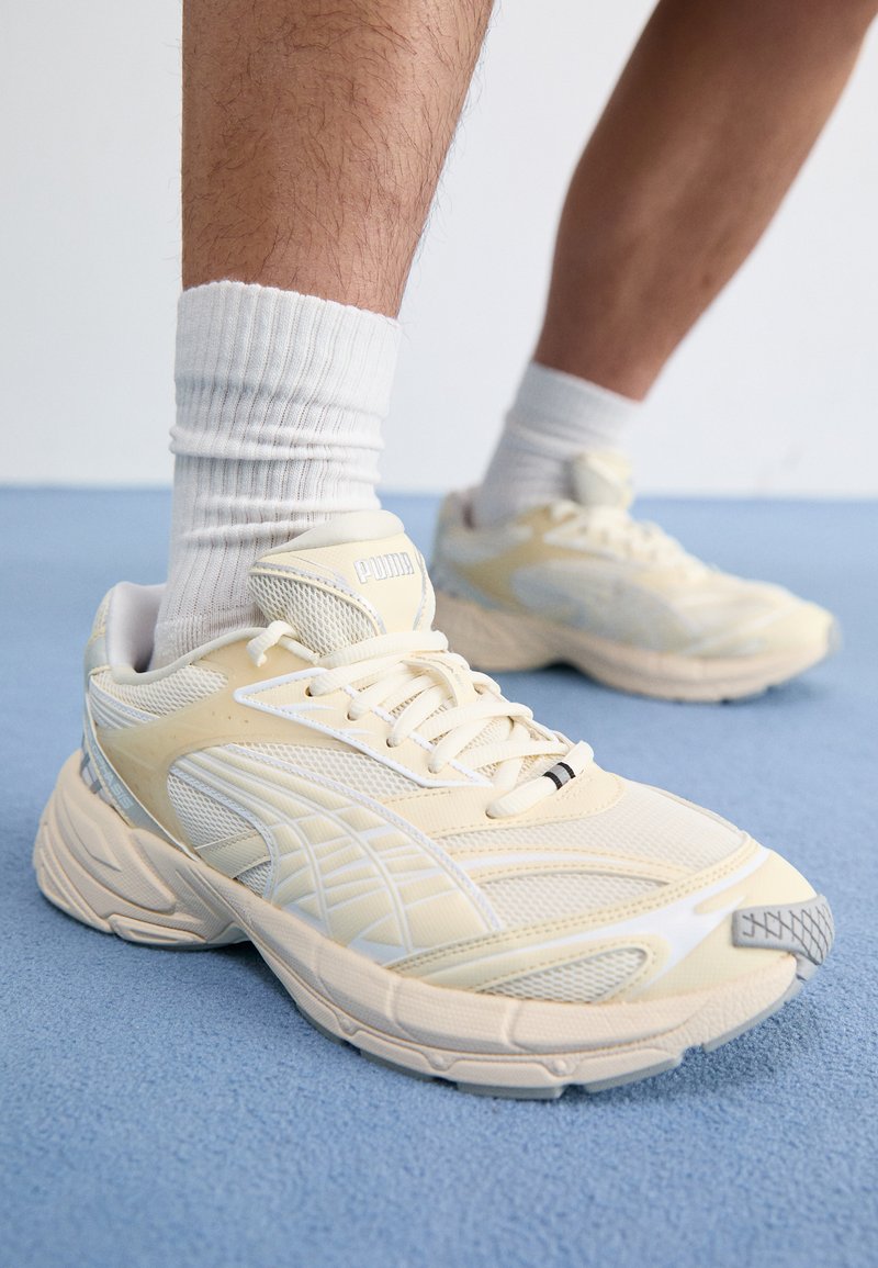 Light yellow athletic shoes with mesh and synthetic overlays, featuring laces, a cushioned sole, and logo on the tongue.