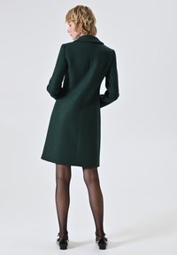 Cappotto verde su misura con una texture liscia, caratterizzato da un colletto a scollo a V e un taglio sopra il ginocchio. Indossato con collant neri e scarpe.
