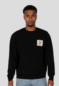 Felpa nera con un piccolo design a forma di quadrato che presenta un personaggio stilizzato. Tessuto morbido, scollatura a girocollo e polsini a coste. Vestibilità casual.