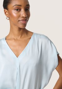 Blusa azzurro chiaro, con texture, scollo a V e maniche corte larghe. Il tessuto morbido si adagia delicatamente, mettendo in mostra una vestibilità rilassata. Orecchini a cerchio in argento.