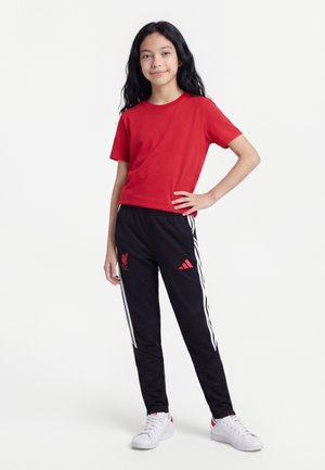 Młoda osoba stoi z ręką na biodrze, ubrana w czerwoną koszulkę, czarne spodnie dresowe Adidas z białymi paskami oraz białe buty sportowe.