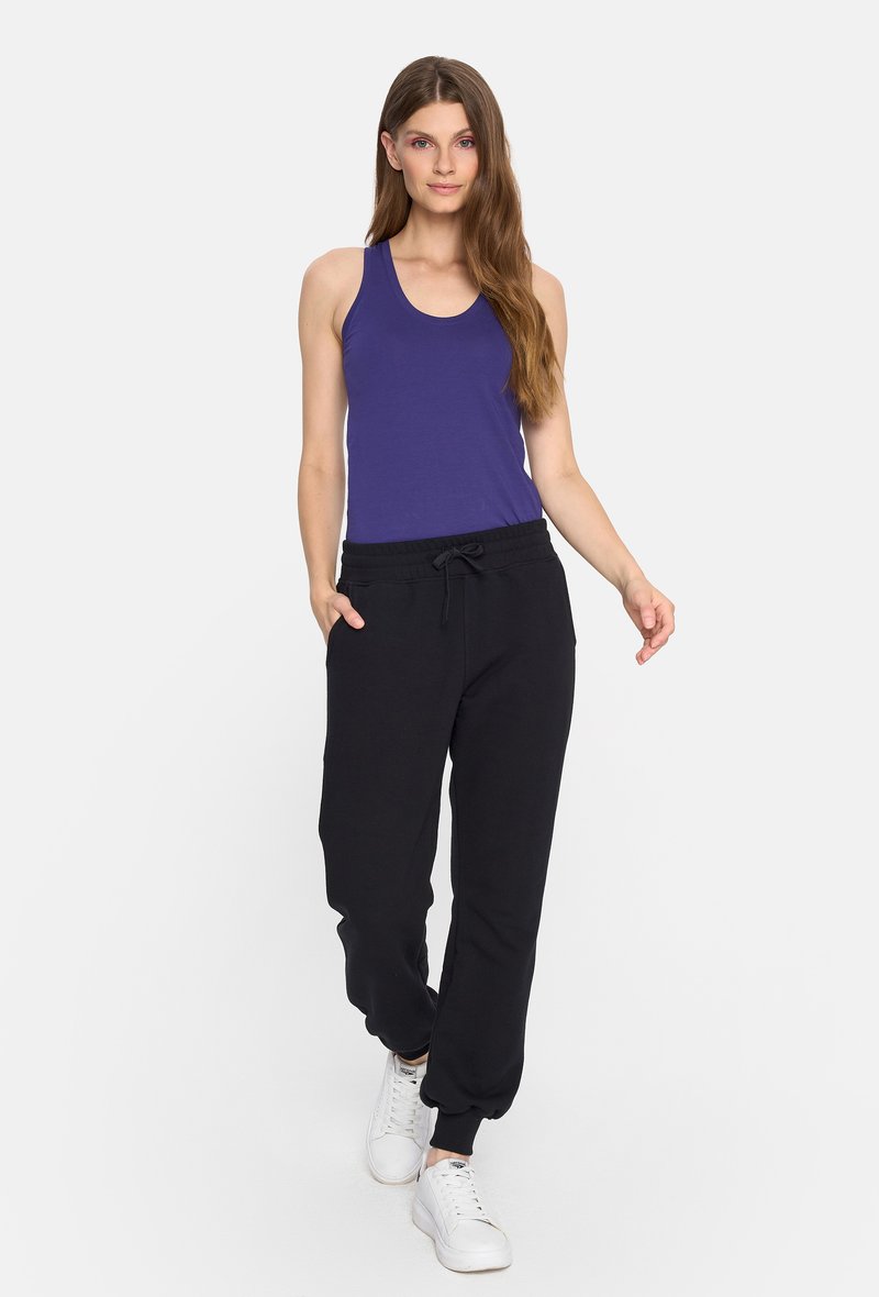Fioletowy top na ramiączkach z okrągłym dekoltem, w zestawieniu z czarnymi spodniami dresowymi z elastycznym pasem i zwężanymi nogawkami. Białe tenisówki.