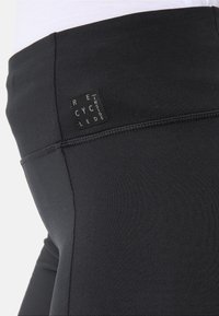 Leggings neri realizzati in materiale elastico con una superficie liscia. Presentano un'etichetta di tessuto riciclato ben visibile e cuciture a piatto.