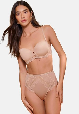 Woman with long dark hair wearing nude-colored high-waist lace lingerie set with bra and matching panties, standing against plain background.