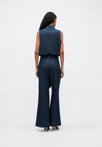 Denim jumpsuit with a sleeveless design, dark blue color, loose-fitting wide legs, and two back pockets. Smooth texture, minimal stitching.