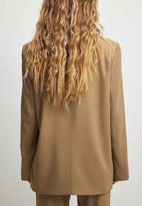 Hnědý dlouhý blazer s přizpůsobeným střihem, jemnou texturou a strukturovanými rameny, pohled zezadu. Jsou vidět dlouhé vlnité vlasy.