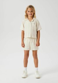 Chemise en coton beige clair à manches courtes avec une poche, assortie à un short à cordon de serrage. Complétée par des baskets blanches et des chaussettes.