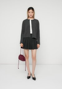 Cardigan en tricot gris à manches longues, associé à un haut blanc et une jupe plissée noire. La tenue comprend un sac pochette bordeaux et des chaussures à talons noirs.