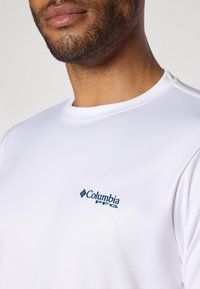 Valkoinen lyhythihainen paita, jossa on pyöreä kaula-aukko, valmistettu sileästä kankaasta. Paidassa on tumma sininen Columbia PFG -logo vasemmassa rinnassa.