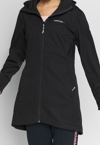 Černá softshellová bunda s kapucí, celoplošným zipem, dvěma bočními kapsami a šedým logem na hrudi. Hladká textura.
