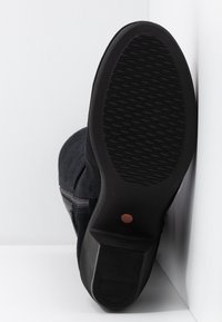 Stivale nero con suola in gomma testurizzata e tacco basso, appoggiato con la suola contro un angolo bianco su una superficie bianca.