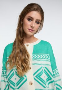 Minkštas kardiganas mėlynai žalsvoje ir kreminėje spalvose, su geometriniais raštais ir sagų uždarymu. Tekstūruotas audinys, laisvas siluetas.