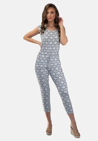 Biały kombinezon z czarnymi geometrycznymi wzorami, krótkie rękawy, dopasowany krój, krótka długość i pasek w talii. Noszony z beżowymi sandałami na koturnie.