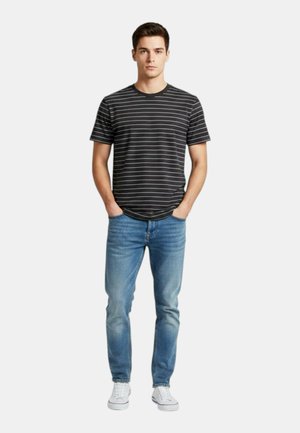 Giovane uomo in piedi che indossa una maglietta a righe bianche e nere, jeans blu, sneakers bianche, con le mani nelle tasche su uno sfondo semplice.