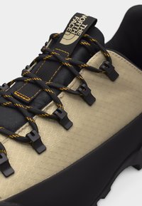 Καφέ και μαύρο παπούτσι πεζοπορίας με μαύρα και κίτρινα κορδόνια, γρήγορα άγκιστρα κορδονιών και λογότυπο «The North Face» στη μαύρη γλώσσα.