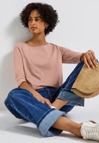 Top de manga larga color rosa claro, hecho de una tela suave, combinado con jeans de pierna ancha azules. Los accesorios incluyen un bolso de ante beige y zapatillas blancas.