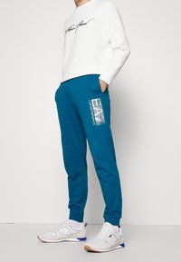 Person som bär en vit sweatshirt och blå joggers med "EA7 Emporio Armani"-logotyp, ihop med vita sneakers och stående mot en vit bakgrund.