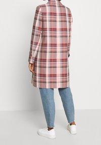 Karierten Mantel in Schattierungen von Rosa, Rot und Blau. Mit langen Ärmeln, einem Kragen und einer taillierten Passform, kombiniert mit hellblauen Jeans und weißen Turnschuhen.