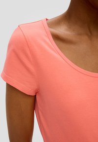 Koraalroze t-shirt met een zachte textuur, ronde halslijn en korte mouwen. Naaddetails zijn zichtbaar op de schouder en de zoom.
