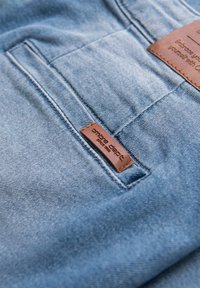 Tissu en denim bleu avec une surface texturée, comportant une étiquette en cuir marron cousue sur la poche arrière, embossée avec "ombre dept."