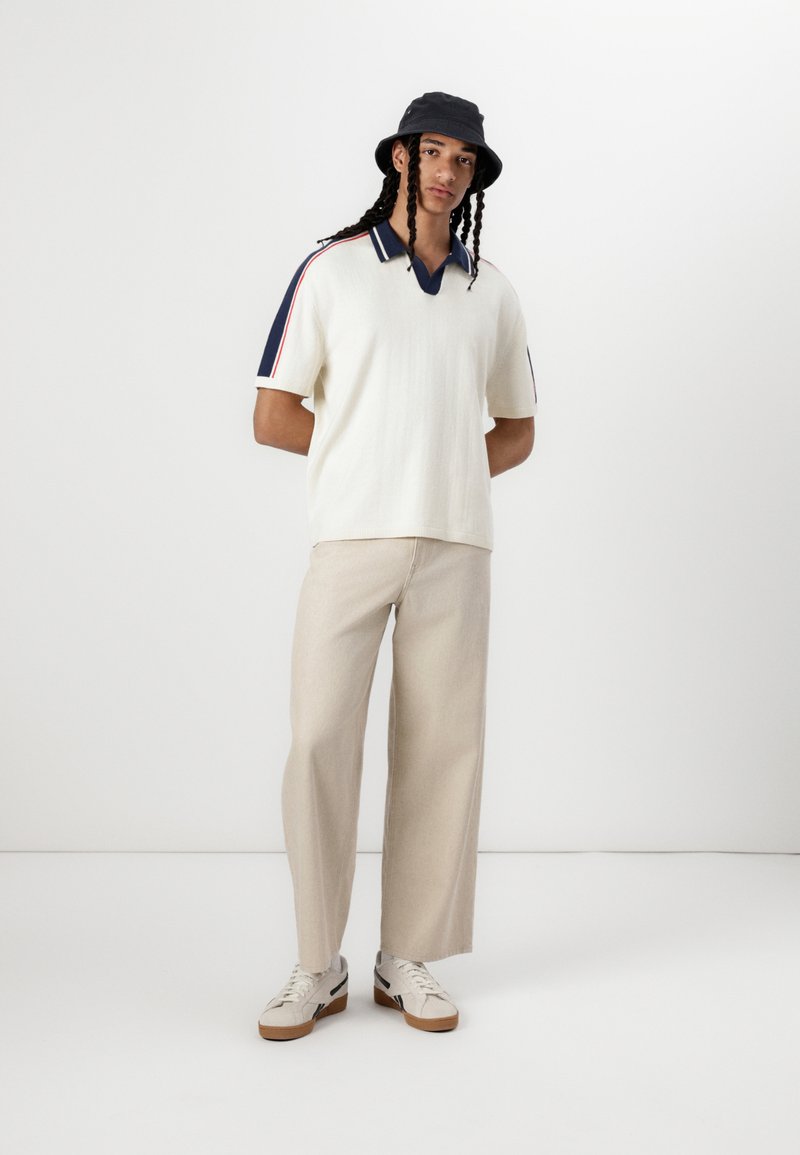 Polo blanco de manga corta con cuello navy y rayas laterales, combinado con pantalones anchos beige y zapatillas neutrales. Sombrero de cubo negro.