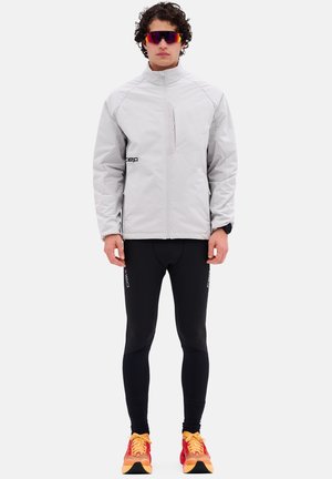 Veste zippée gris clair avec poignets élastiques et poche latérale, portée avec des leggings noirs et des chaussures de course multicolores.