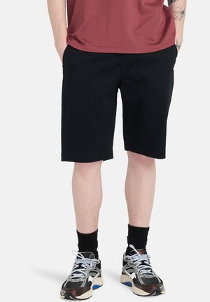 Osoba ubrana w czarne szorty do kolan, czarne skarpetki, czarno-białe buty sportowe i bordową koszulkę, z rękami w kieszeniach.