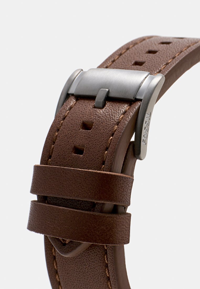 Brązowy skórzany pasek do zegarka z teksturowaną powierzchnią, metalowym zapięciem z grawerunkiem oraz wieloma otworami do regulacji rozmiaru.