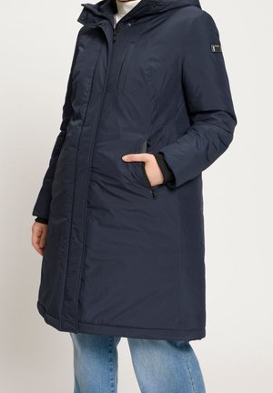 Personne portant un long manteau matelassé bleu marine avec poches zippées et capuche, associé à un jean bleu et un col roulé blanc.