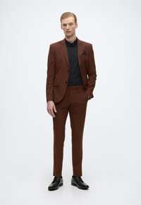 Bruin op maat gemaakt pak met een zwart overhemd, slim fit broek en zwarte schoenen. Kenmerkt zich door een enkel knoops blazer en een pochet detail.