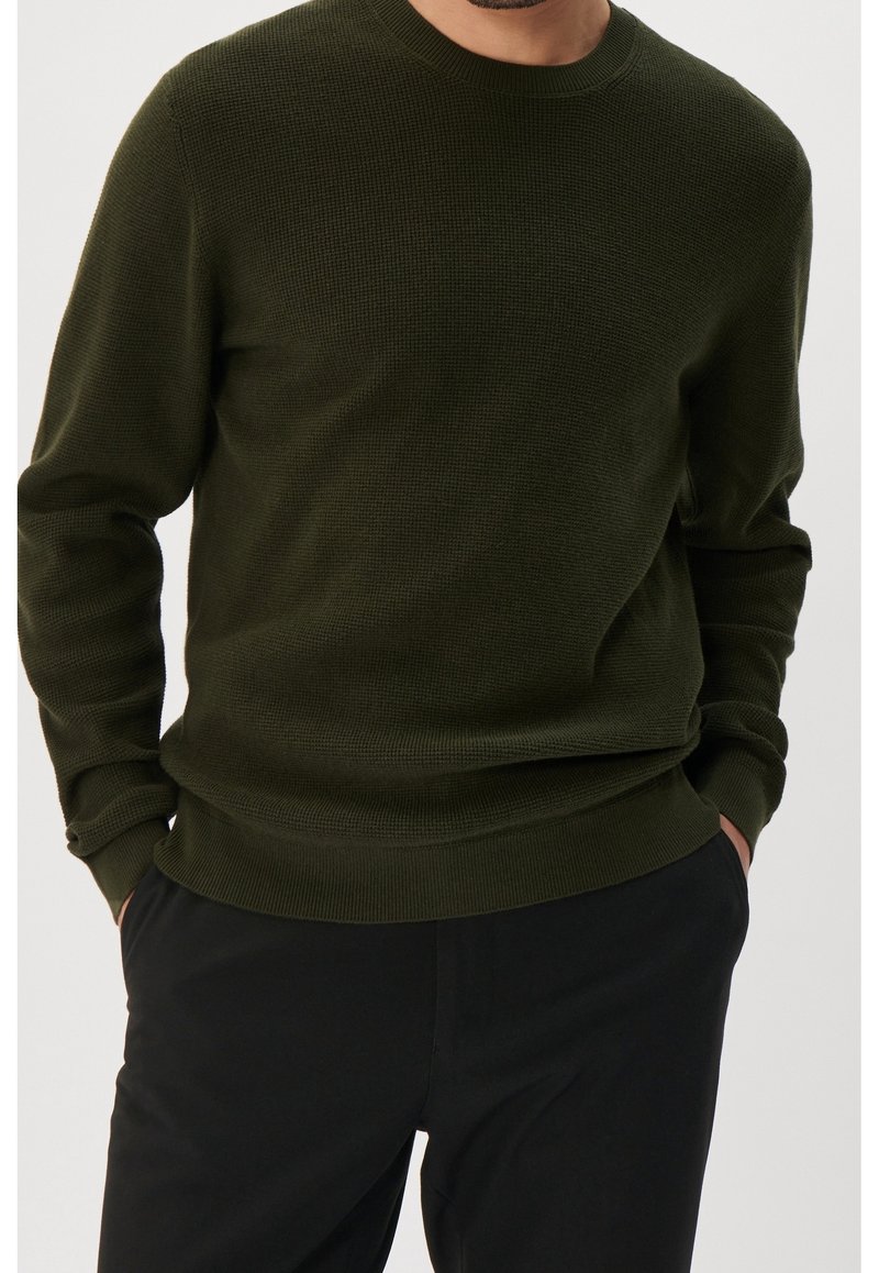 Homme portant un pull en maille texturée vert foncé avec poignets et ourlet côtelés, mains dans les poches, associé à un pantalon noir.
