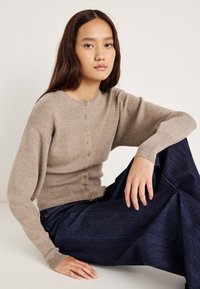 Ανοιχτό μπεζ πουλόβερ με κουμπιά και στρογγυλή λαιμόκοψη, ριμπ μανίκια και εφαρμοστό μέσο, σε συνδυασμό με σκούρα μπλε παντελόνια φαρδιάς γραμμής.