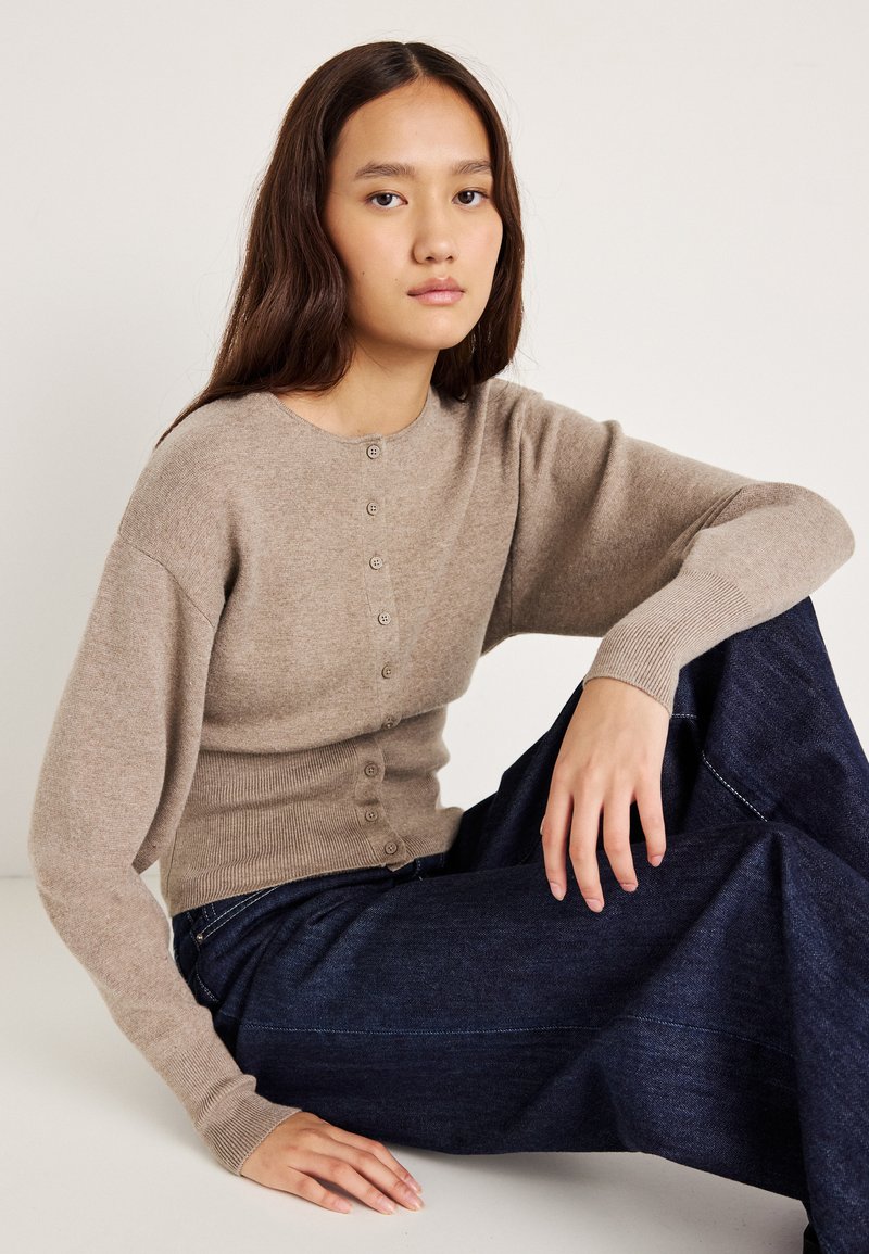 Ανοιχτό μπεζ πουλόβερ με κουμπιά και στρογγυλή λαιμόκοψη, ριμπ μανίκια και εφαρμοστό μέσο, σε συνδυασμό με σκούρα μπλε παντελόνια φαρδιάς γραμμής.