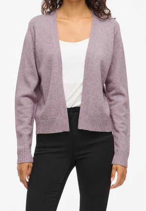 Femme portant un cardigan violet clair ouvert sur un haut blanc et un pantalon noir, debout, les bras détendus le long du corps.