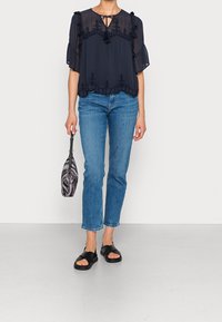 Blusa blu navy con maniche arricciate e dettagli ricamati, abbinata a jeans blu chiaro e sandali neri, mentre tiene una borsa a righe.