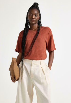 Kvinde med langt fletning hår iført en rustfarvet kortærmet top og hvide højtaljede bukser, der holder en tan clutch i den ene hånd.