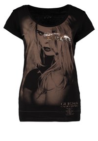 T-shirt noir à manches courtes avec une impression photographique délavée, des accents en sequins et le logo "GUESS" en bas. Texture de tissu douce.