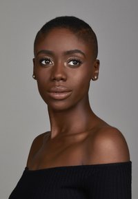 Portrait d'une personne avec les cheveux noirs courts, portant un haut noir à épaules dénudées, avec une peau lisse et un maquillage subtil aux accents dorés.
