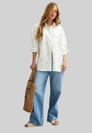 Chemise en coton blanche avec broderie florale jaune, coupe ample, devant à boutons. Associée à un jean en denim large bleu clair et à un sac fourre-tout en osier.