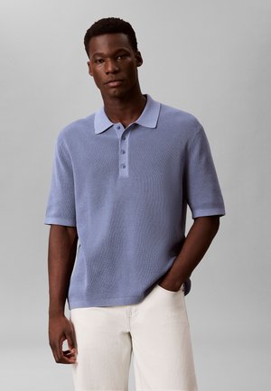 Mężczyzna ubrany w jasnoniebieską, teksturowaną koszulkę polo z krótkim rękawem oraz białe spodnie, stojący z jedną ręką w kieszeni na jednolitym szarym tle.