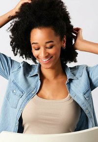 Camicia di denim di colore azzurro chiaro, con due tasche sul petto, indossata sopra un top beige. Il soggetto ha i capelli ricci e sta interagendo con i capelli.
