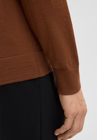 Eine Nahaufnahme einer Hand, die einen braunen Pullover trägt, der das strukturierte Gewebe und den gerippten Saum zeigt, neben dunklen Hosen.