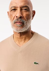 Beige v-ringad tröja gjord av ett texturerat stickat material, med en liten grön alligatorlogga på bröstet. Rent och enkelt design.