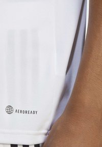 Maglietta atletica bianca realizzata in tessuto testurizzato, con un logo nero "AEROREADY" sull'orlo e strisce vicino al fondo.
