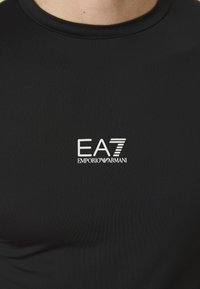 Svart treningst-skjorte laget av teksturert stoff. Har en hvit EA7 Emporio Armani-logo på forsiden. Crew-neck design.