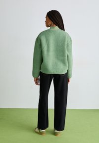 Fuzzy green jacket with a textured finish, high collar, paired with black wide-leg trousers and green accents on footwear.