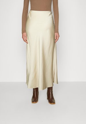 Femme portant une jupe midi beige en satin, un haut marron à manches longues et des bottes à talons en cuir marron, debout devant un fond uni.